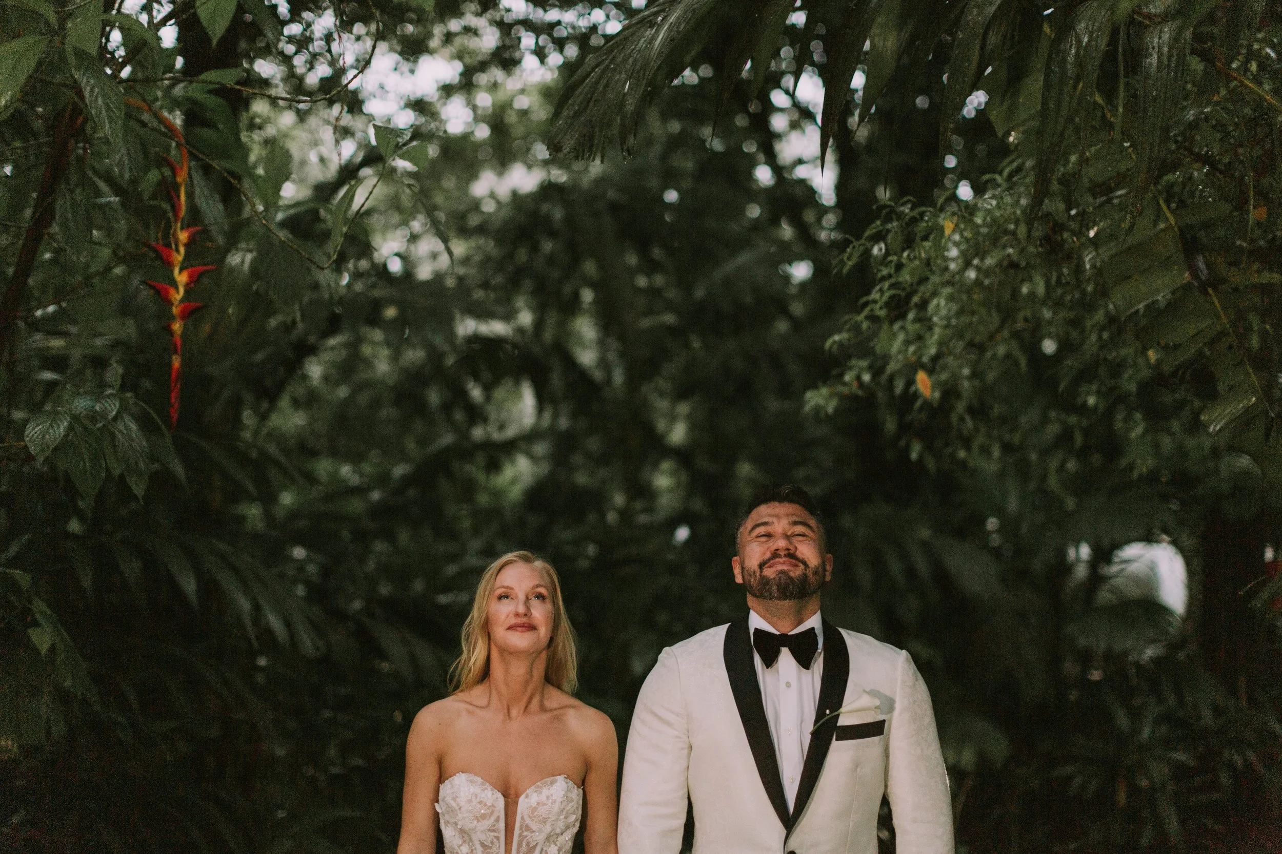 Bride and groom in a white shawl-lapel tuxedo standing under dense rainforest foliage with a red heliconia bloom