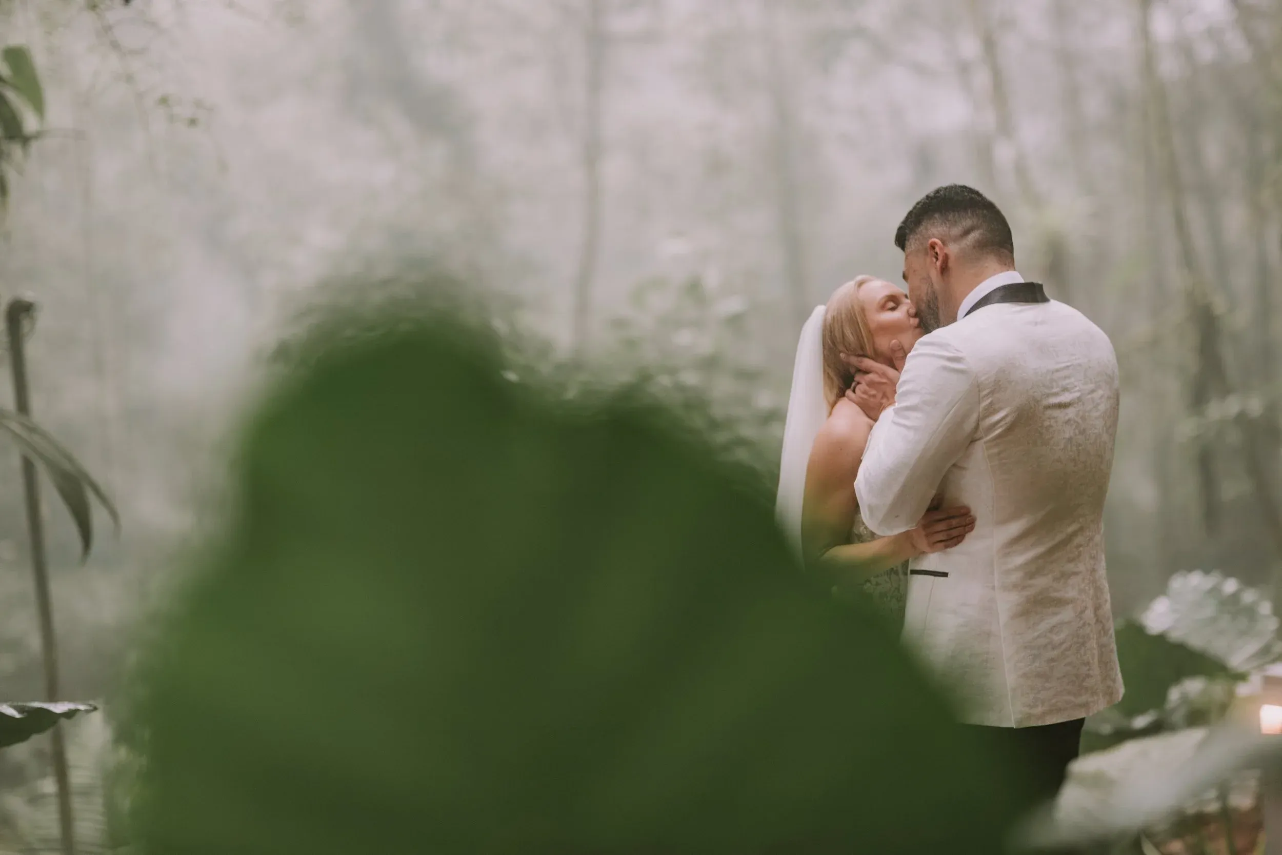 Bride and groom kiss beneath fog in a forest, his white jacquard dinner jacket against the green