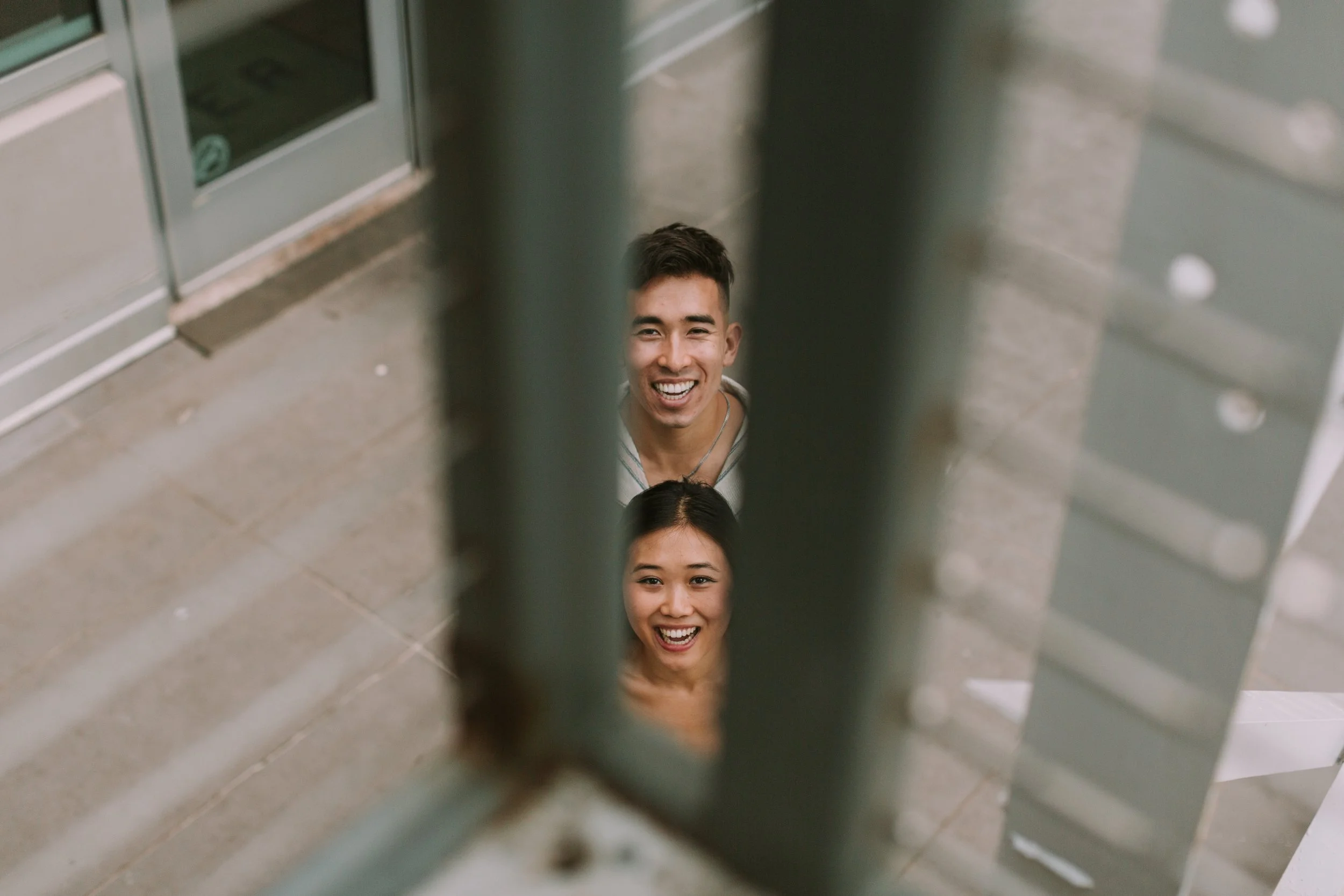 Ben and Grace grinning up at the camera through the slats of a metal railing on an Austin sidewalk