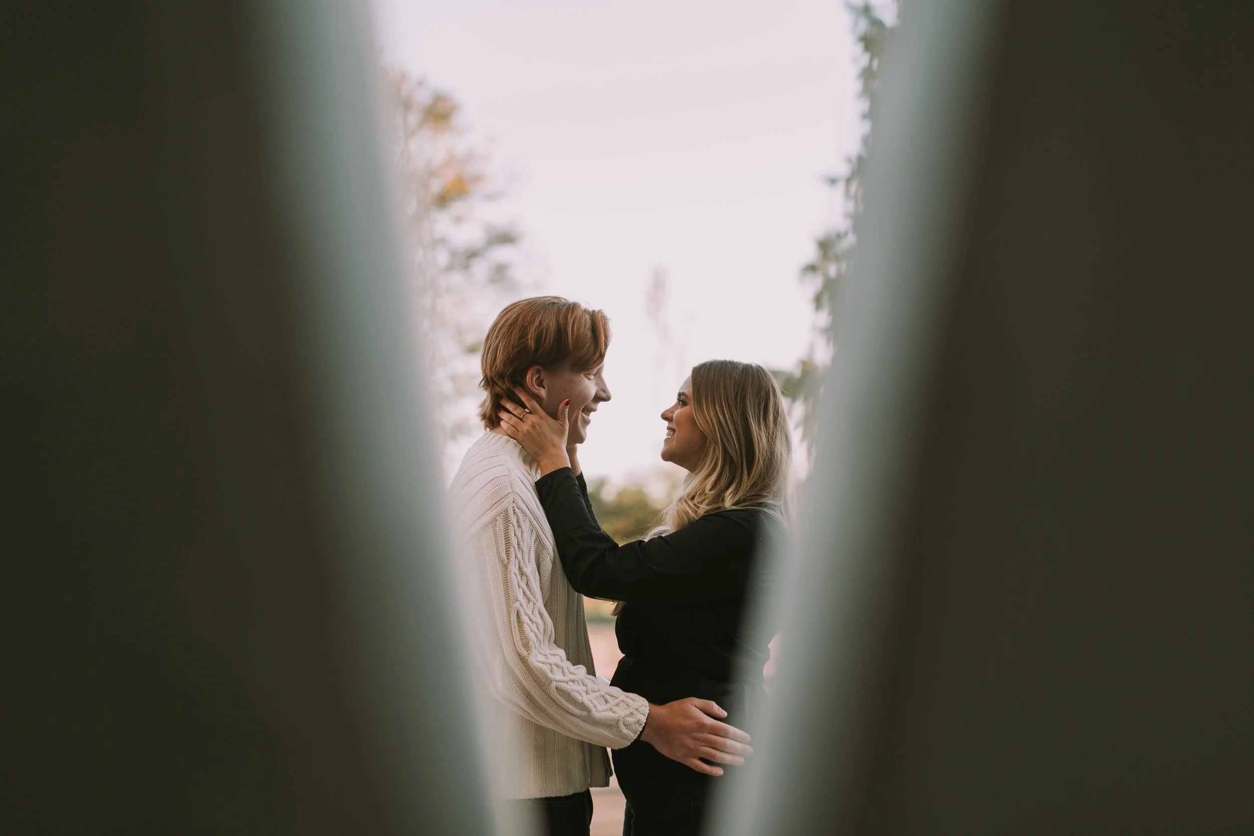 Riley holding Jake's face as they laugh together at dusk in Austin's Mueller neighborhood, framed between blurred posts
