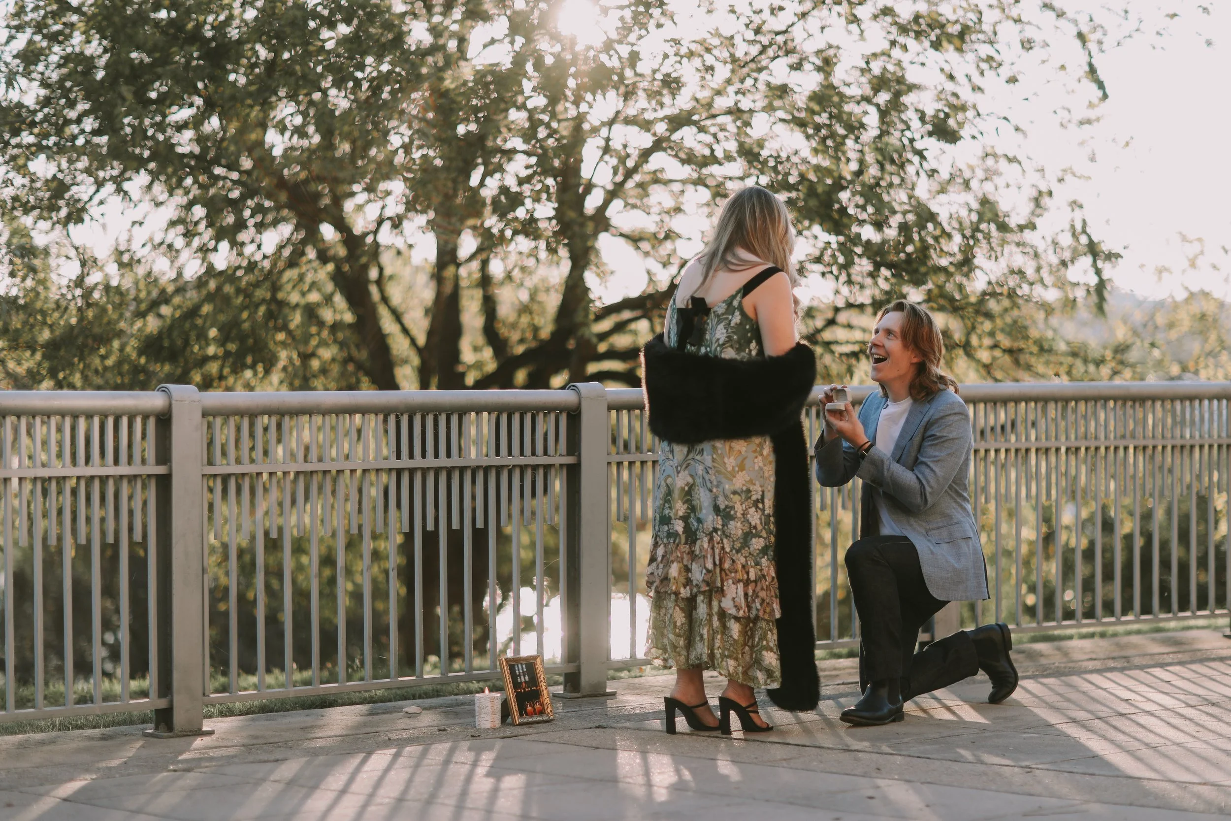 Jake on one knee at a riverside walkway railing in Austin, Riley in a floral dress reacting, golden-hour sun