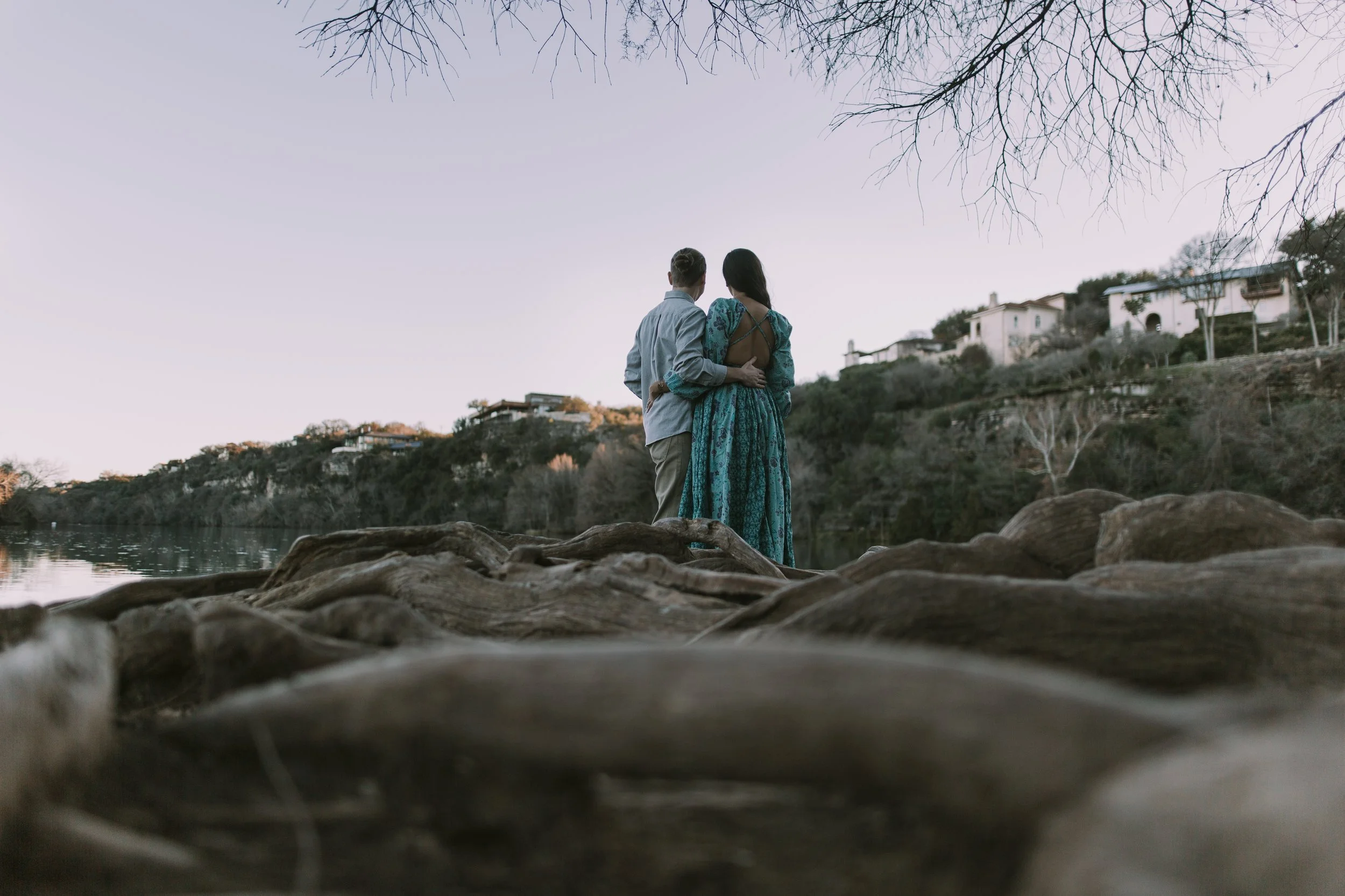 Couple embracing on tangled driftwood at the water's edge at dusk, hillside homes across the lake