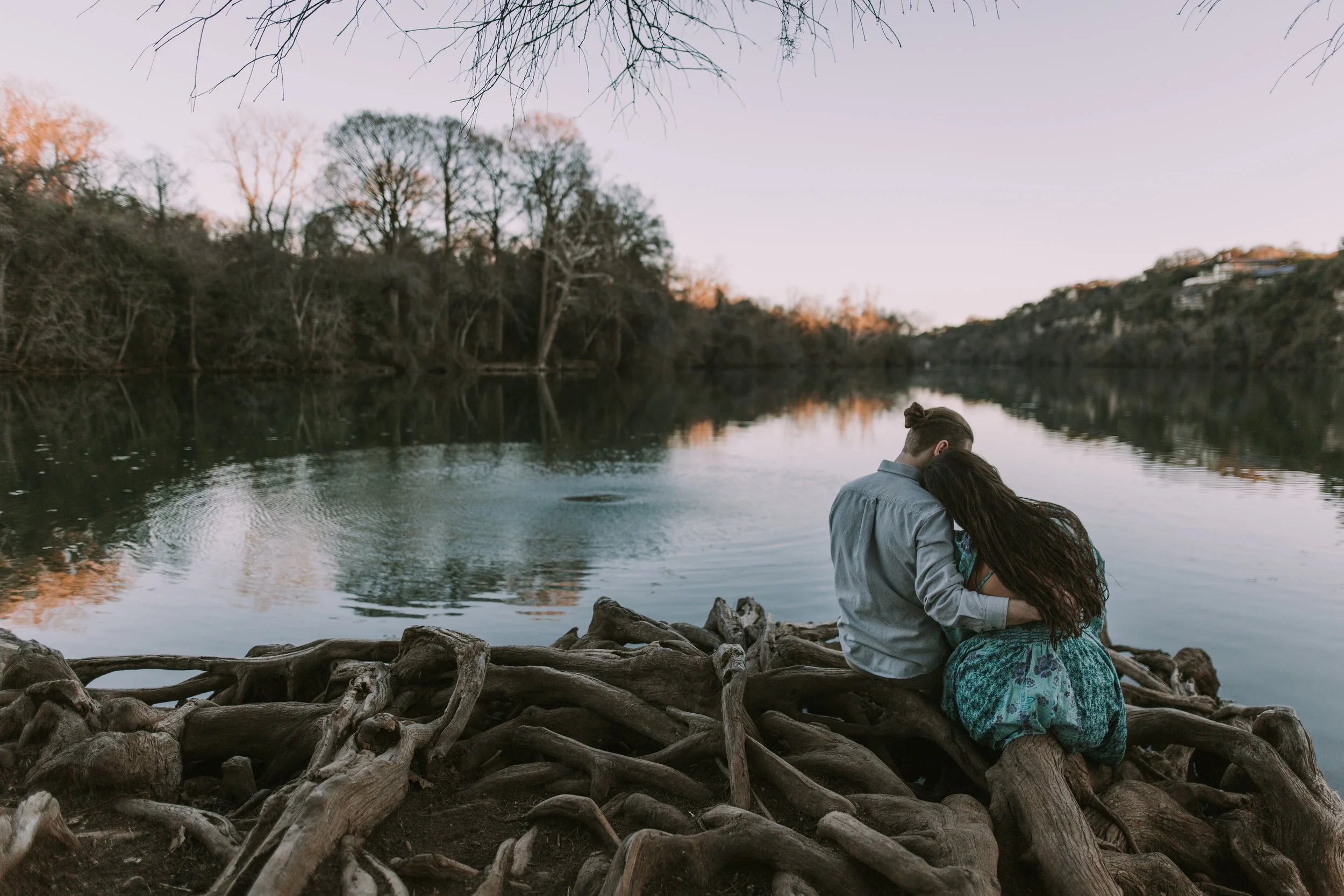 Jasmin resting her head on Grant's shoulder, seated on driftwood by the calm water in soft evening light
