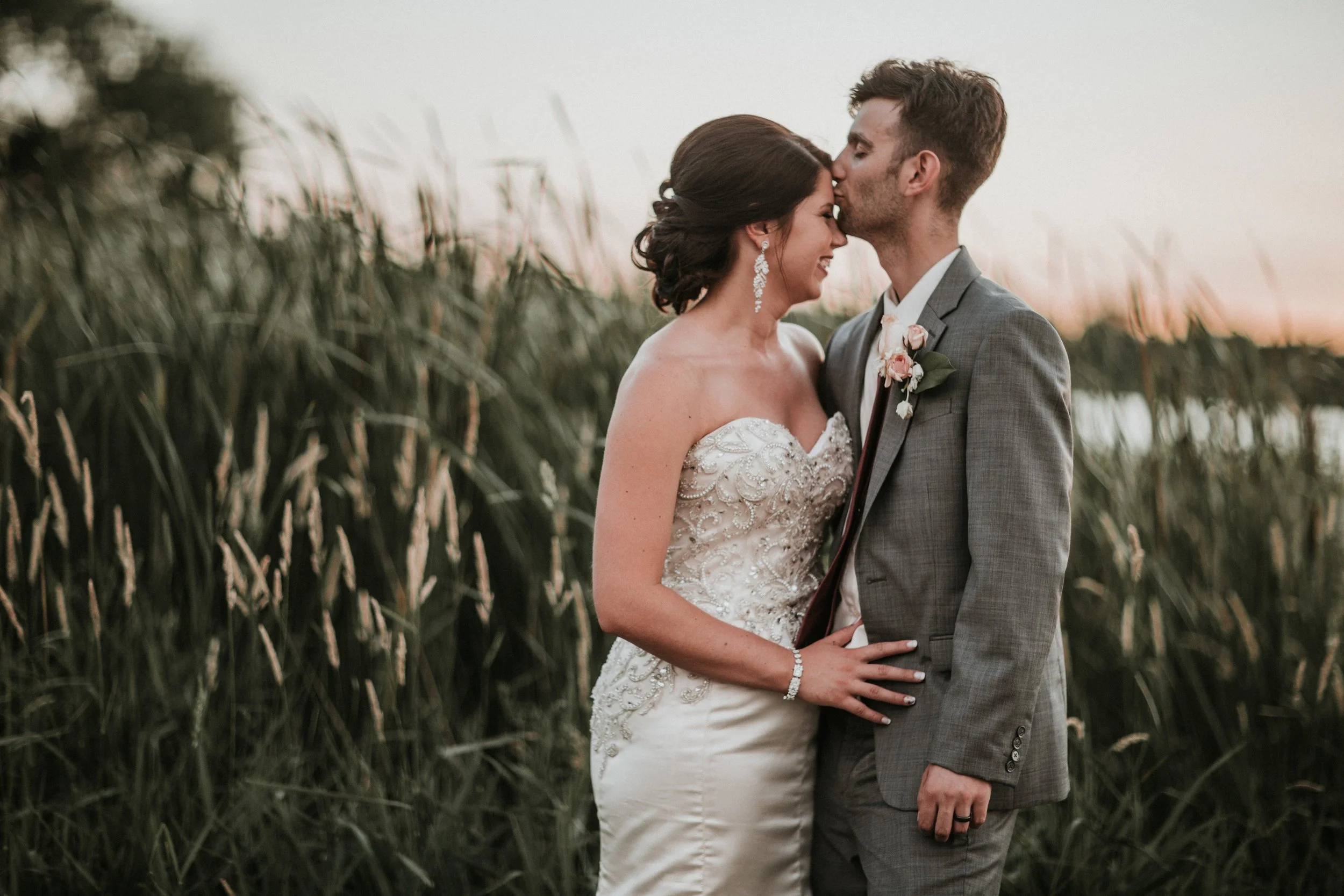 Groom kisses the bride's temple in waist-high golden grass at last light, still water behind them