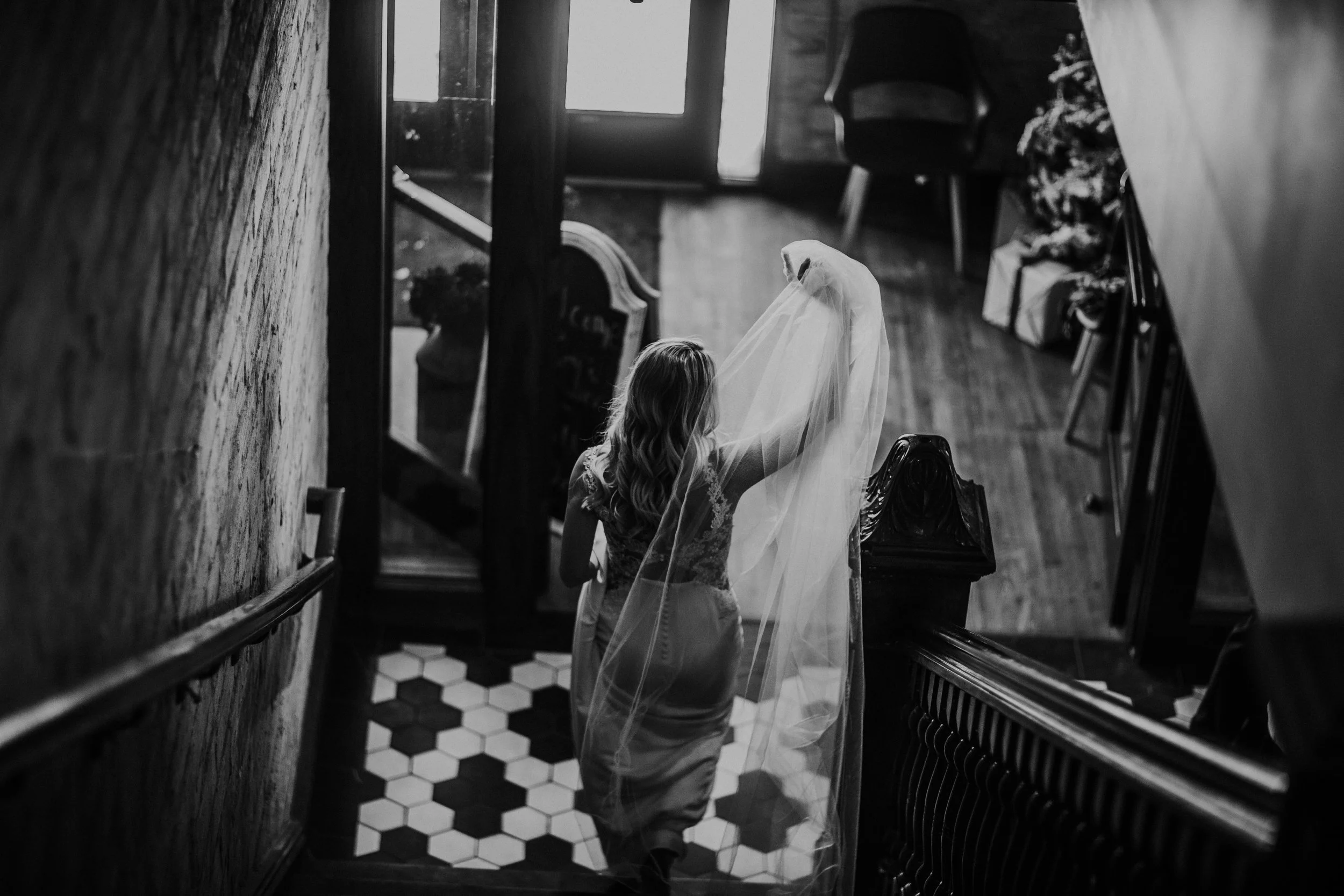 Bride carries her veil down a staircase to a hex-tile landing, in black and white