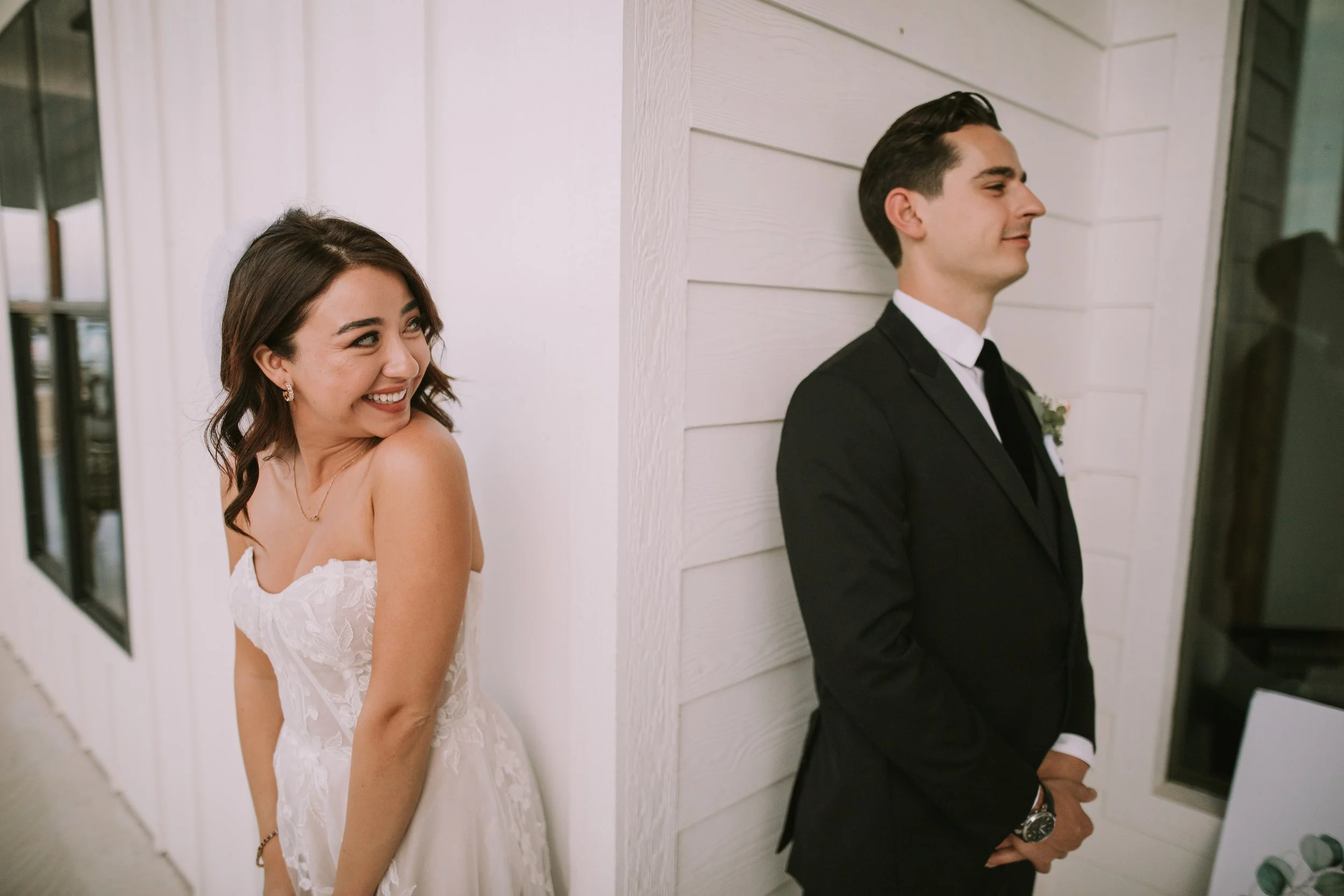 Bride laughs around the corner from her groom just before their first look, white farmhouse wall