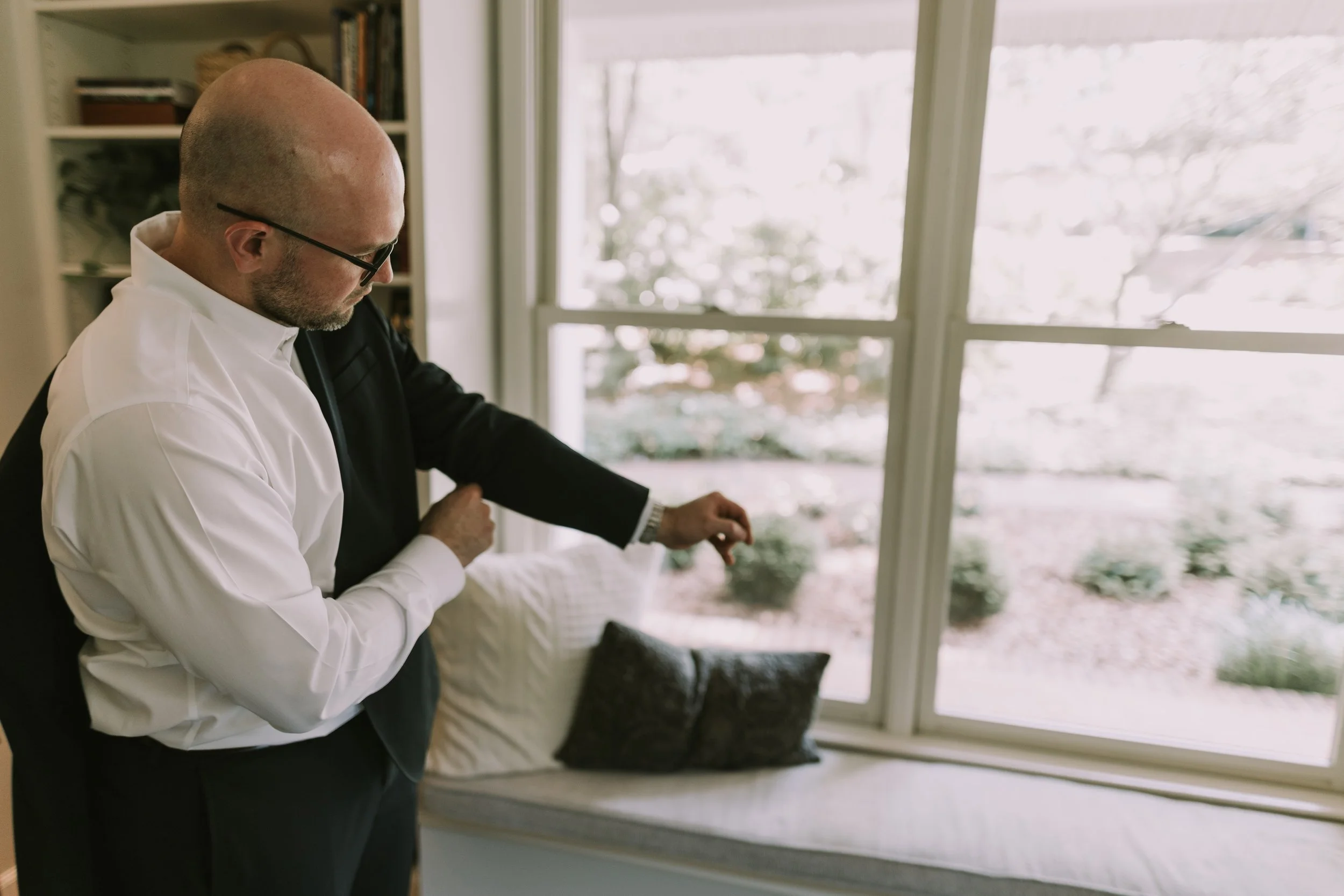 Groom pulling on his suit jacket beside a bright window before his Charlotte wedding
