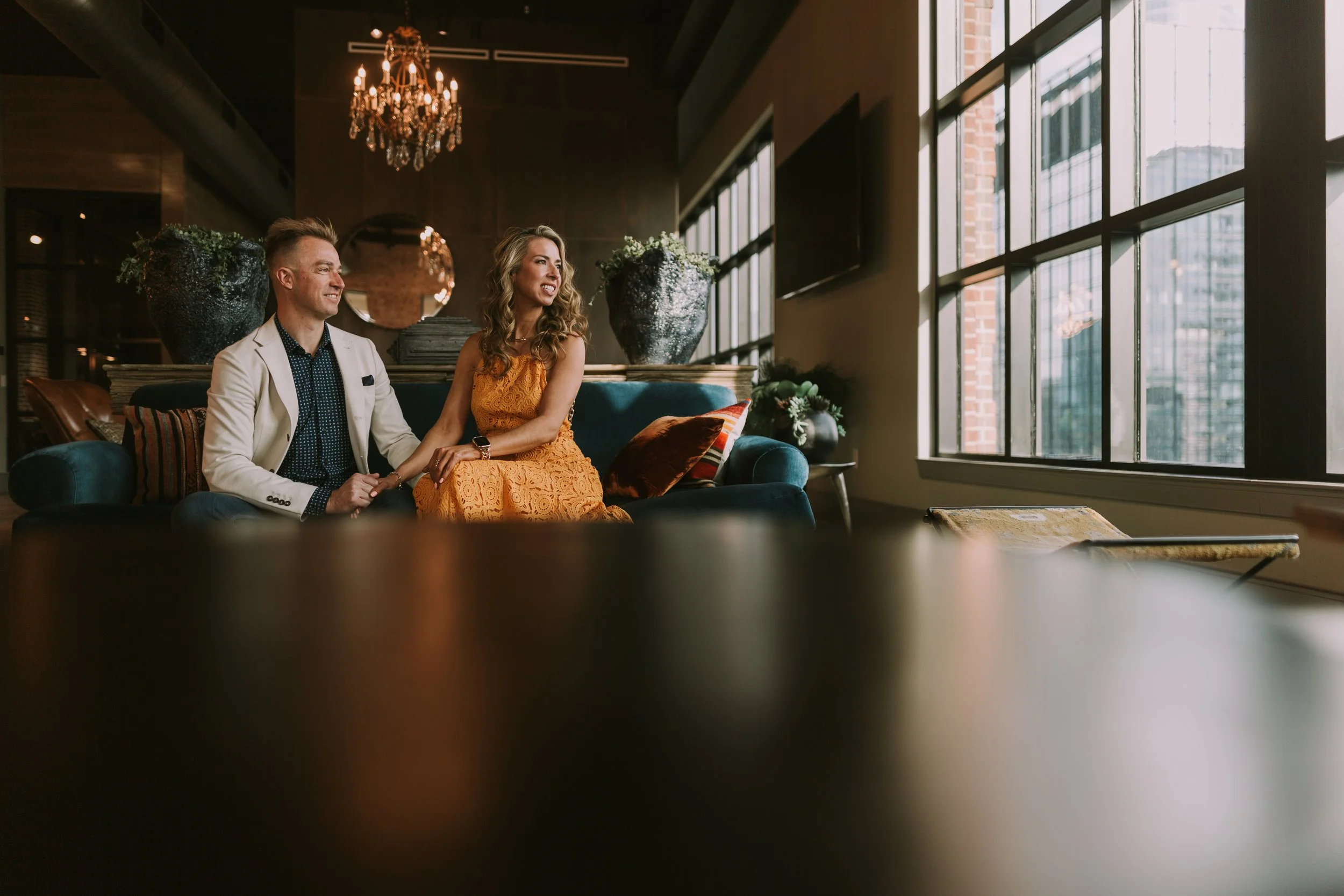 Couple holding hands on a teal velvet sofa in an industrial loft lounge with downtown Austin outside the windows