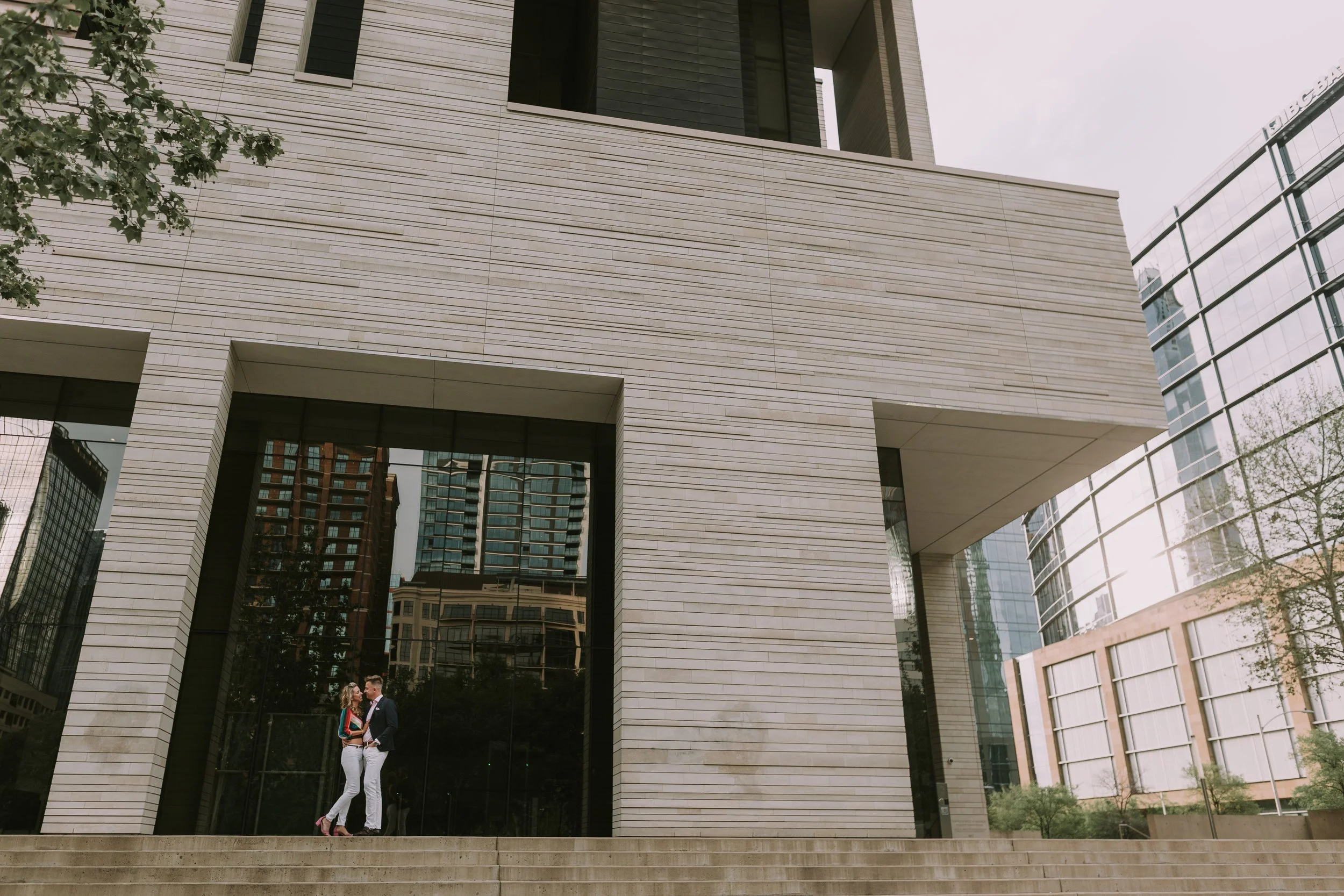 Mathias and Doralicia embracing on a downtown Austin plaza, small against a limestone tower reflecting the skyline