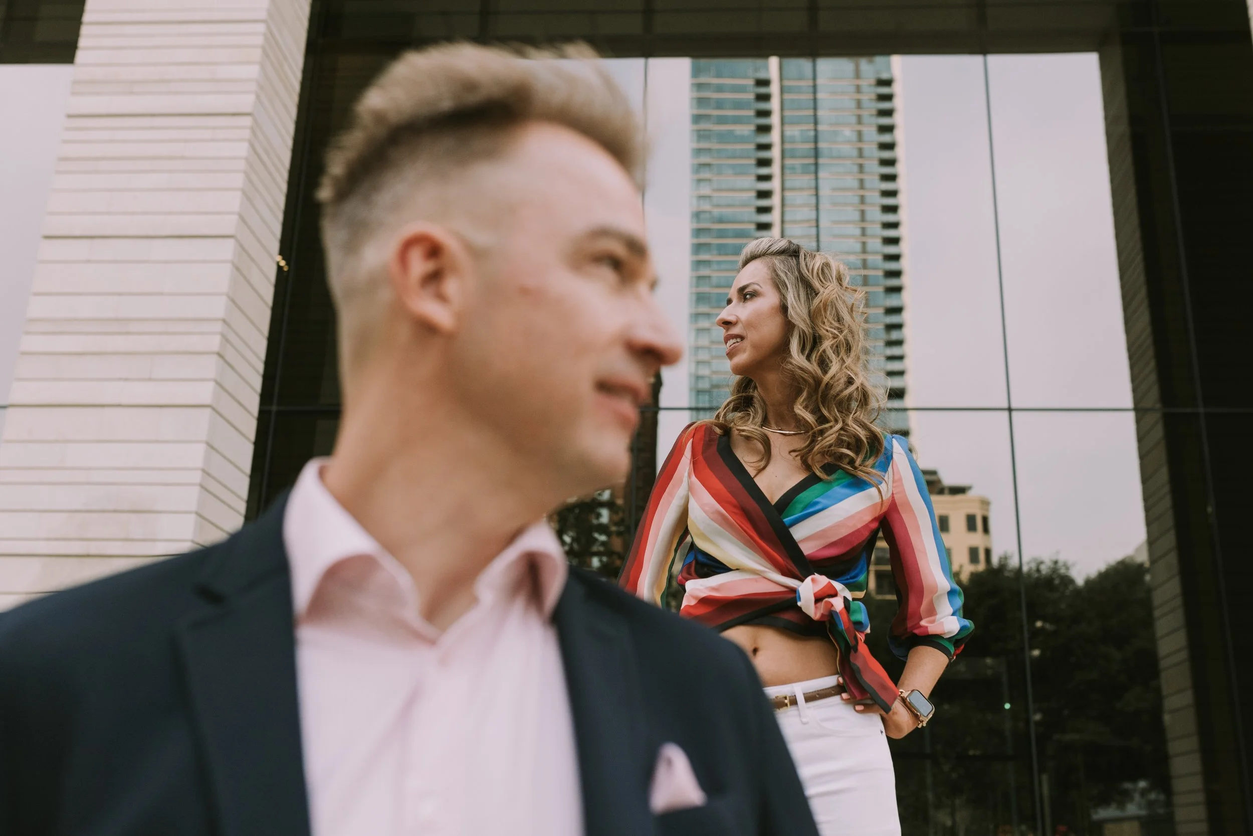 Doralicia in a rainbow-striped top before a reflective glass tower, Mathias blurred in the foreground
