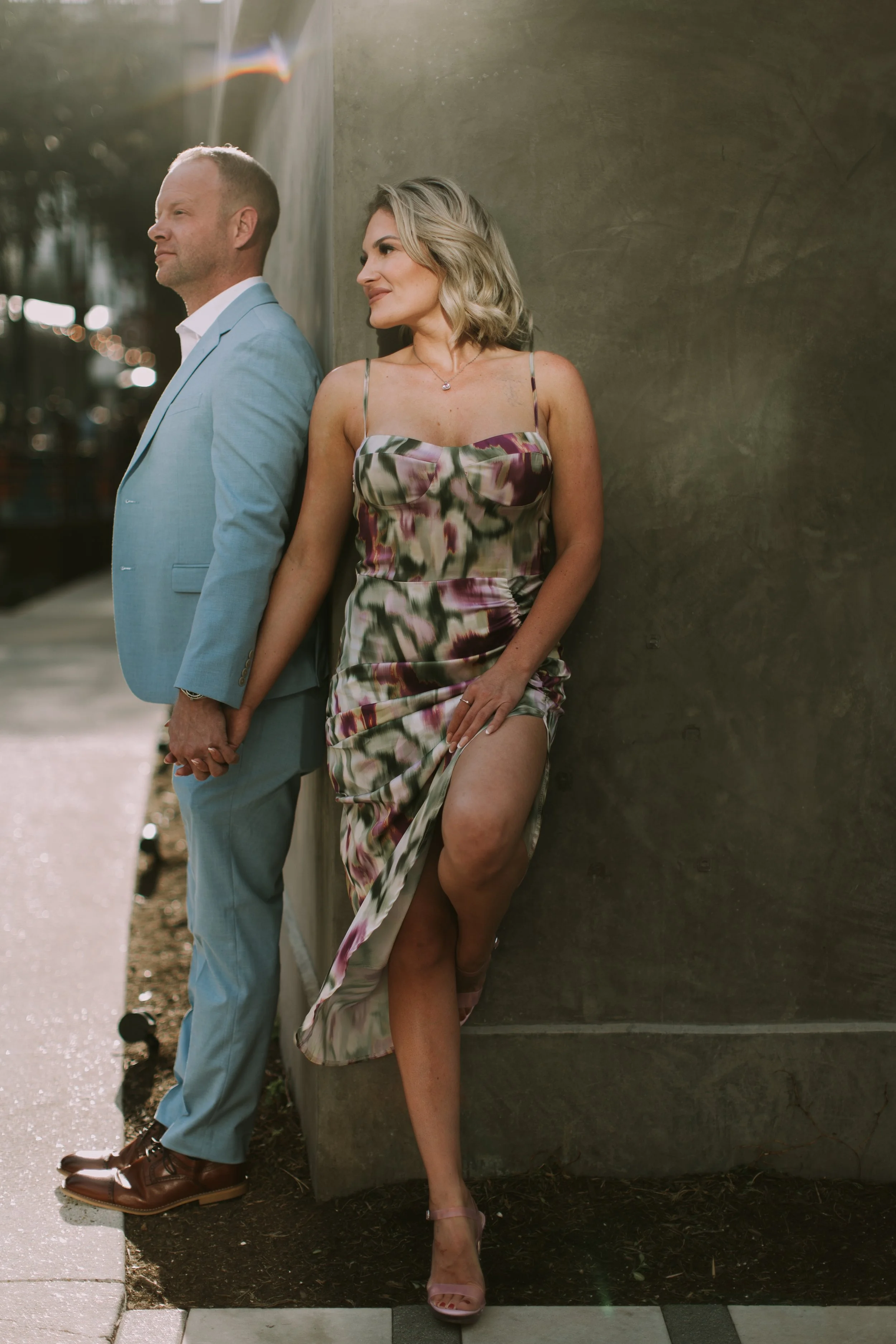 Ryan in a light-blue suit holding hands with Tessa in a floral slip dress against a dark wall in warm Austin sun