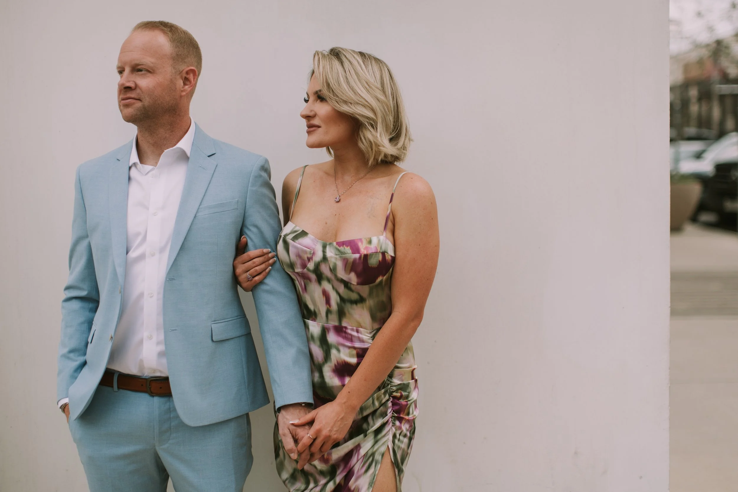 Ryan and Tessa arm in arm against a white wall in Austin, pale blue suit and floral slip dress, gazing the same way