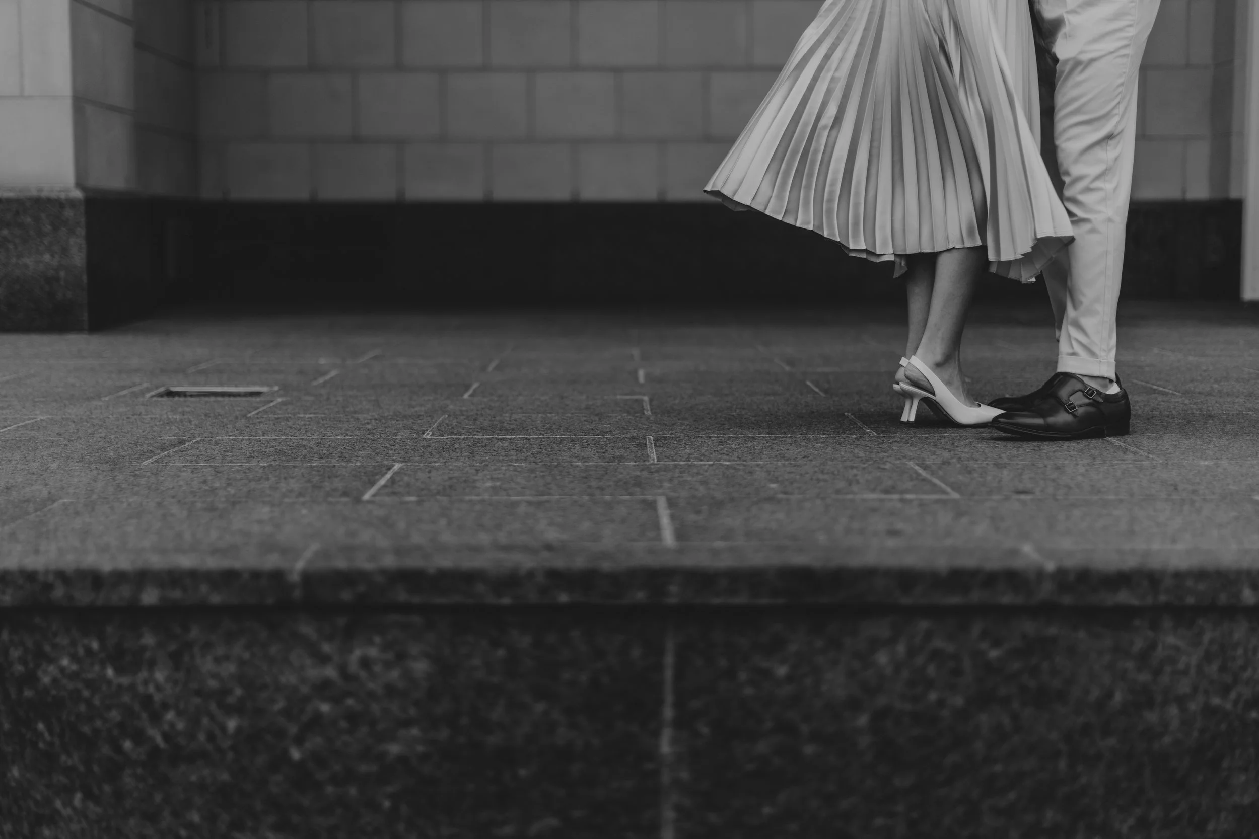 Black and white detail of a couple's feet, pleated skirt and slingback heels beside leather dress shoes