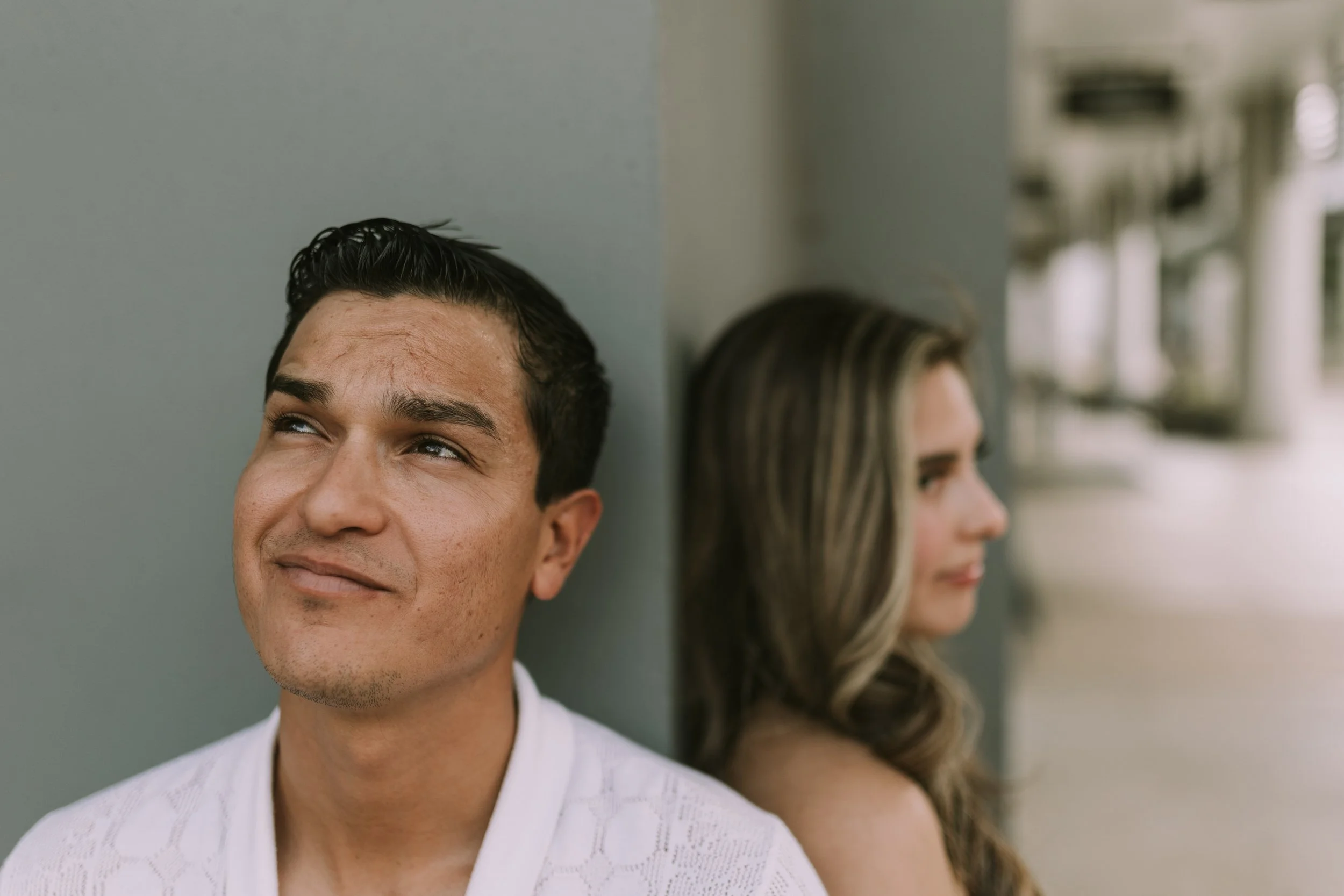 Eduardo gazing up beside a sage-grey column in downtown Austin, Wendy soft-focus on the far side