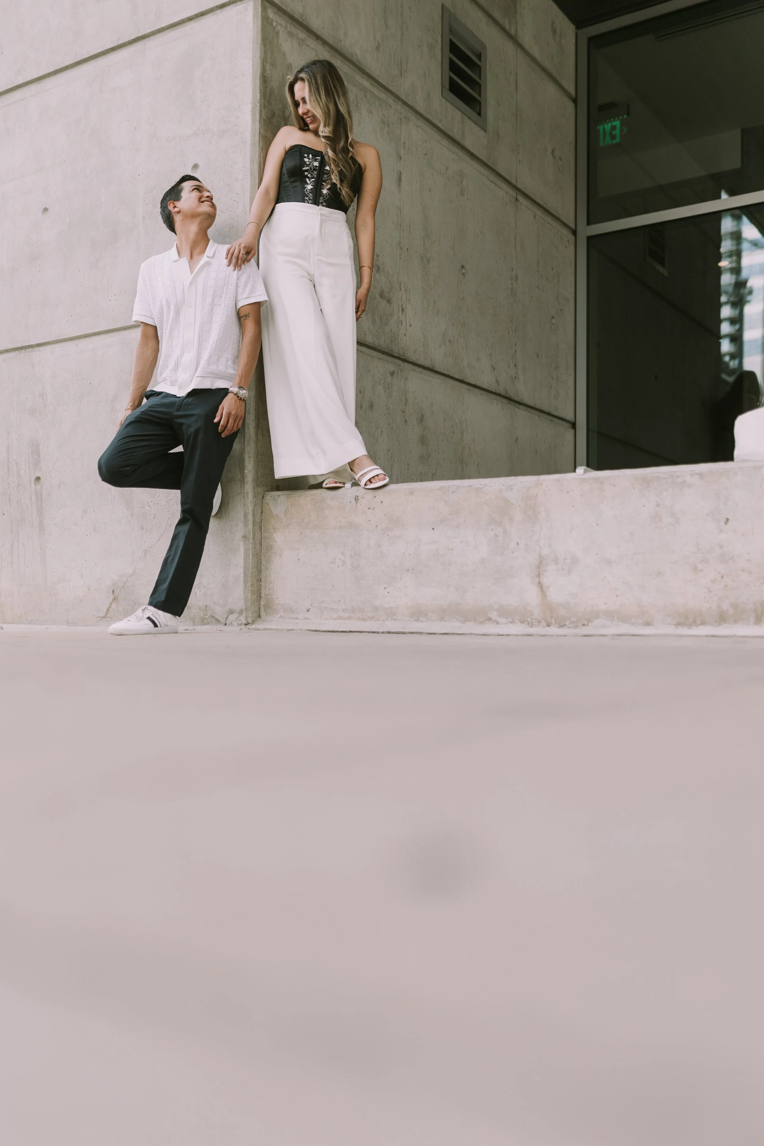 Wendy in a black corset and white pants on a concrete ledge smiling down at Eduardo in the Seaholm District, Austin