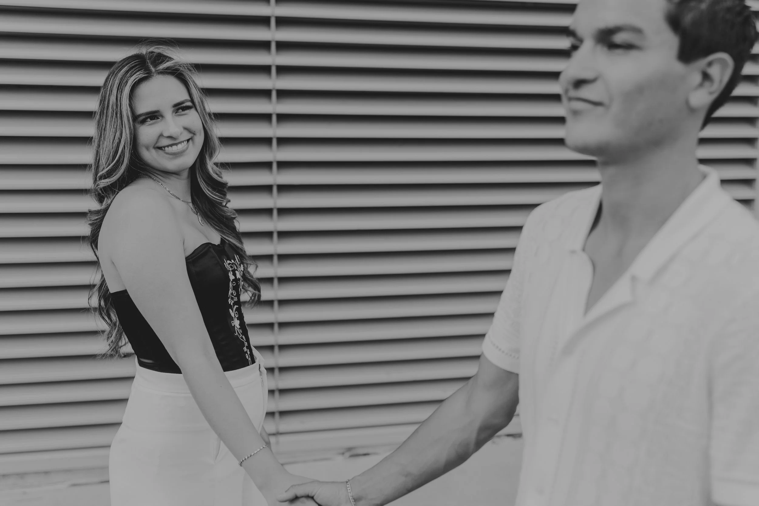 Black and white frame of Wendy smiling over her shoulder, holding Eduardo's hand before a louvered metal wall