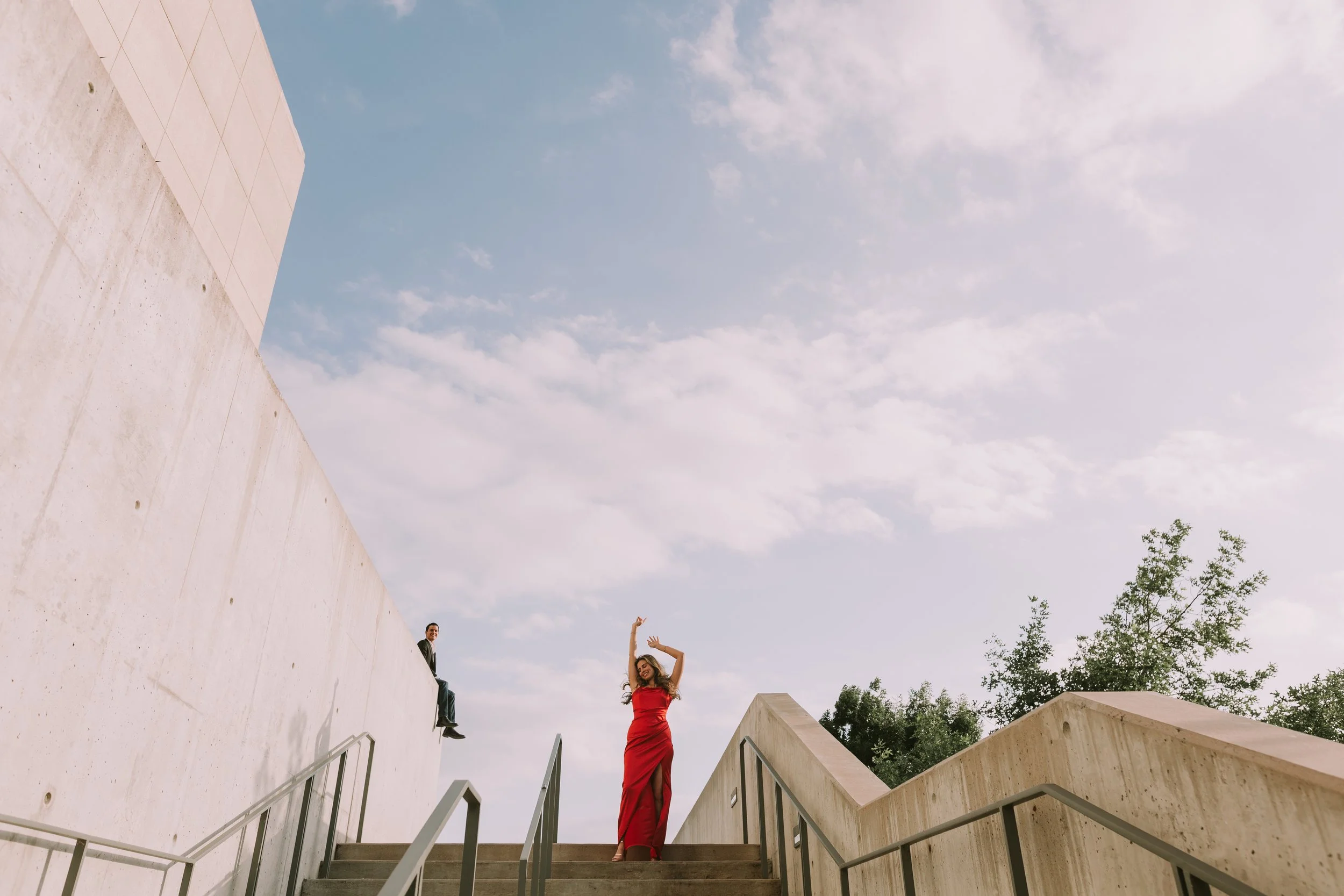 She dances in a red dress on brutalist concrete stairs in Austin while he watches from the wall above